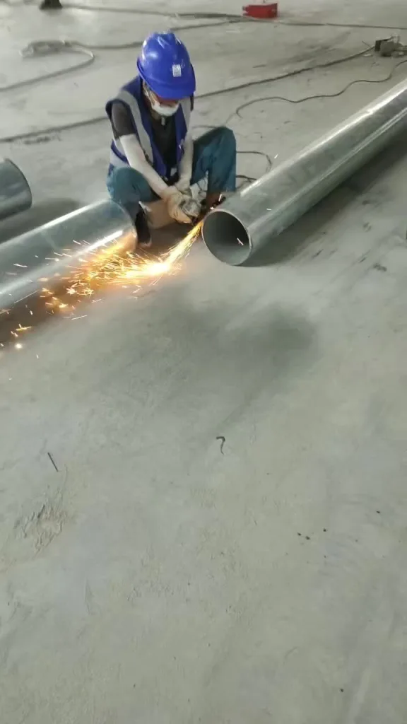 A welder grinding a steel pipe on the shop floor — properly fitted gloves let the hands move freely through grinding, fitting, and welding.